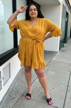 Load image into Gallery viewer, Full-body front view of a size 20 Boohoo mustard yellow faux wrap mini dress with white polka dots, a ruffle hem, and a subtle bell sleeve styled with black heels on a size 18/20 model.
