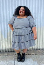Load image into Gallery viewer, Additional full-body front view of a size 28 Simply Be brand black and white gingham plaid mini-print tiered prairie dress with black trim and puff sleeves styled with black combat boots on a size 24/26 model. The photo is taken outside in natural lighting.
